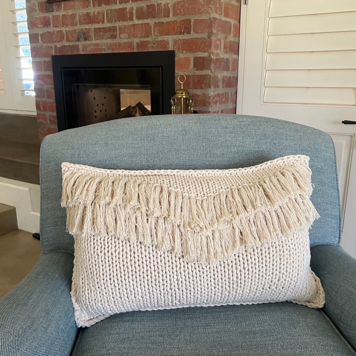 A cream-colored knit lumbar pillow with tiered fringe, resting on a blue-gray armchair in front of a brick fireplace.