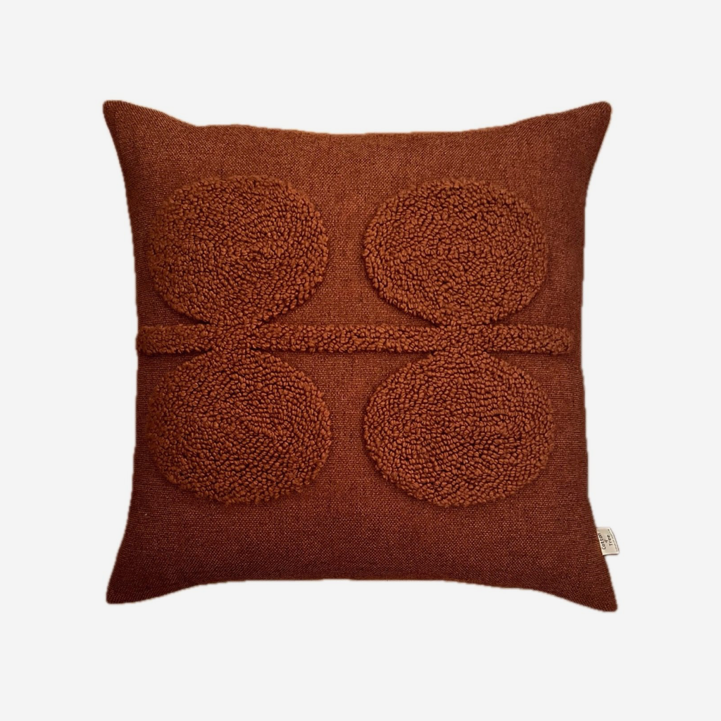 A square rust-brown textured throw pillow featuring four raised circular tufted motifs and a horizontal textured stripe across the center, shown on a light gray background.