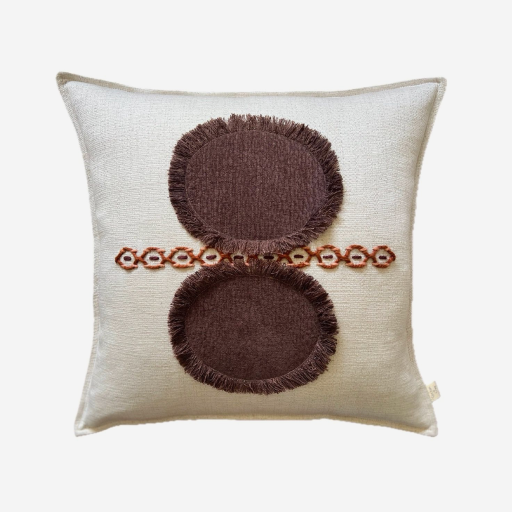 A square off-white throw pillow featuring a brown embroidered design with two circular motifs and a horizontal line of small floral accents.