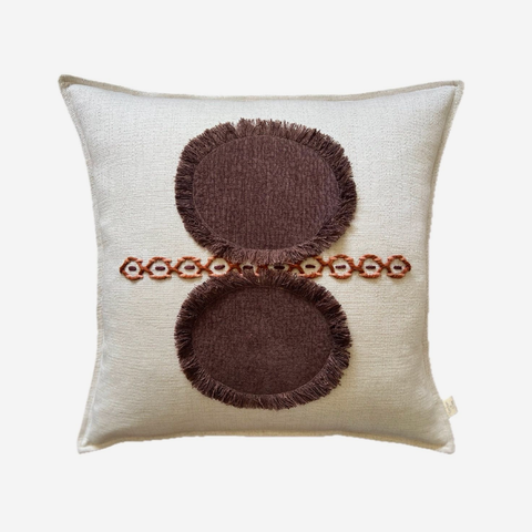 A square off-white throw pillow featuring a brown embroidered design with two circular motifs and a horizontal line of small floral accents.