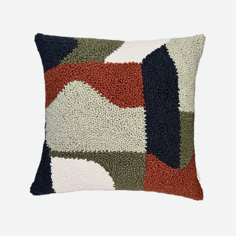 A square throw pillow featuring a textured, chunky abstract geometric pattern in navy blue, rust orange, olive green, cream, and off-white, set against a plain light gray background.