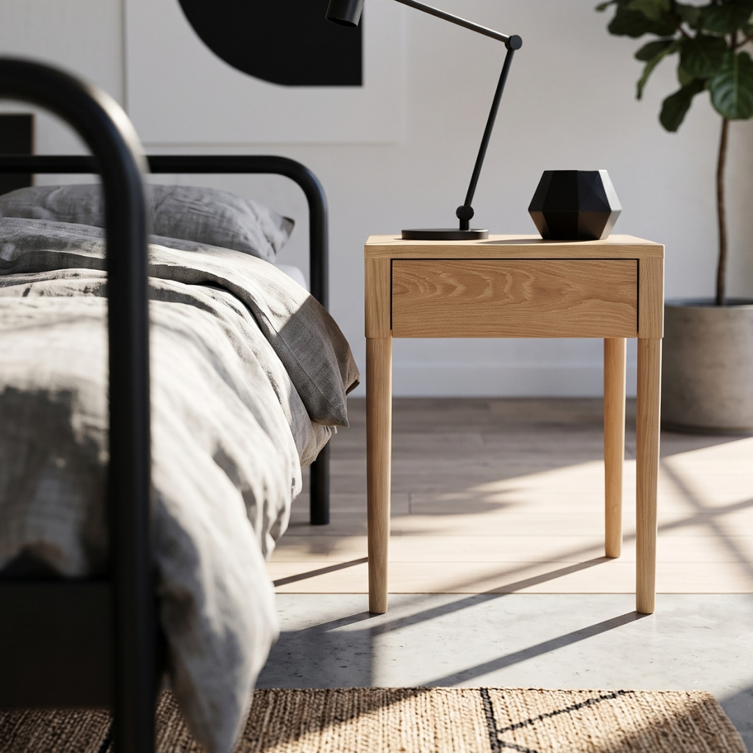 A light natural wood nightstand with a single drawer, styled in a modern bedroom next to a bed with gray bedding. A black desk lamp and a faceted black vase sit on top of the nightstand, with warm sunlight casting shadows on the floor.