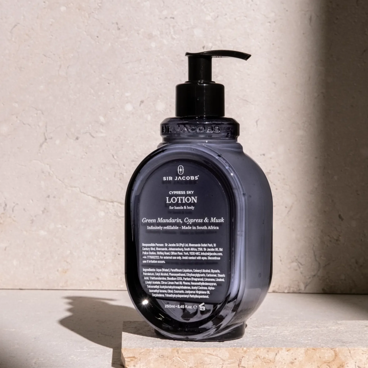 A bottle of Sir Jacobs Cyprime Soy Lotion in a dark, translucent smoky gray bottle with a black pump dispenser, set on a stone slab against a textured beige stone wall.