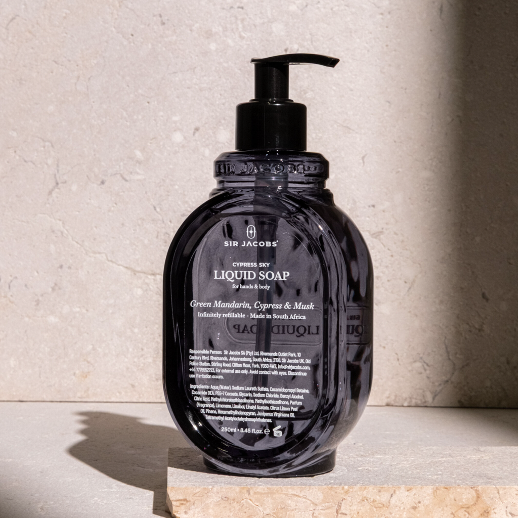 A bottle of Sir Jacobs Green Mandarin, Cypress & Musk liquid soap with a black pump dispenser, set on a stone slab against a textured stone wall.